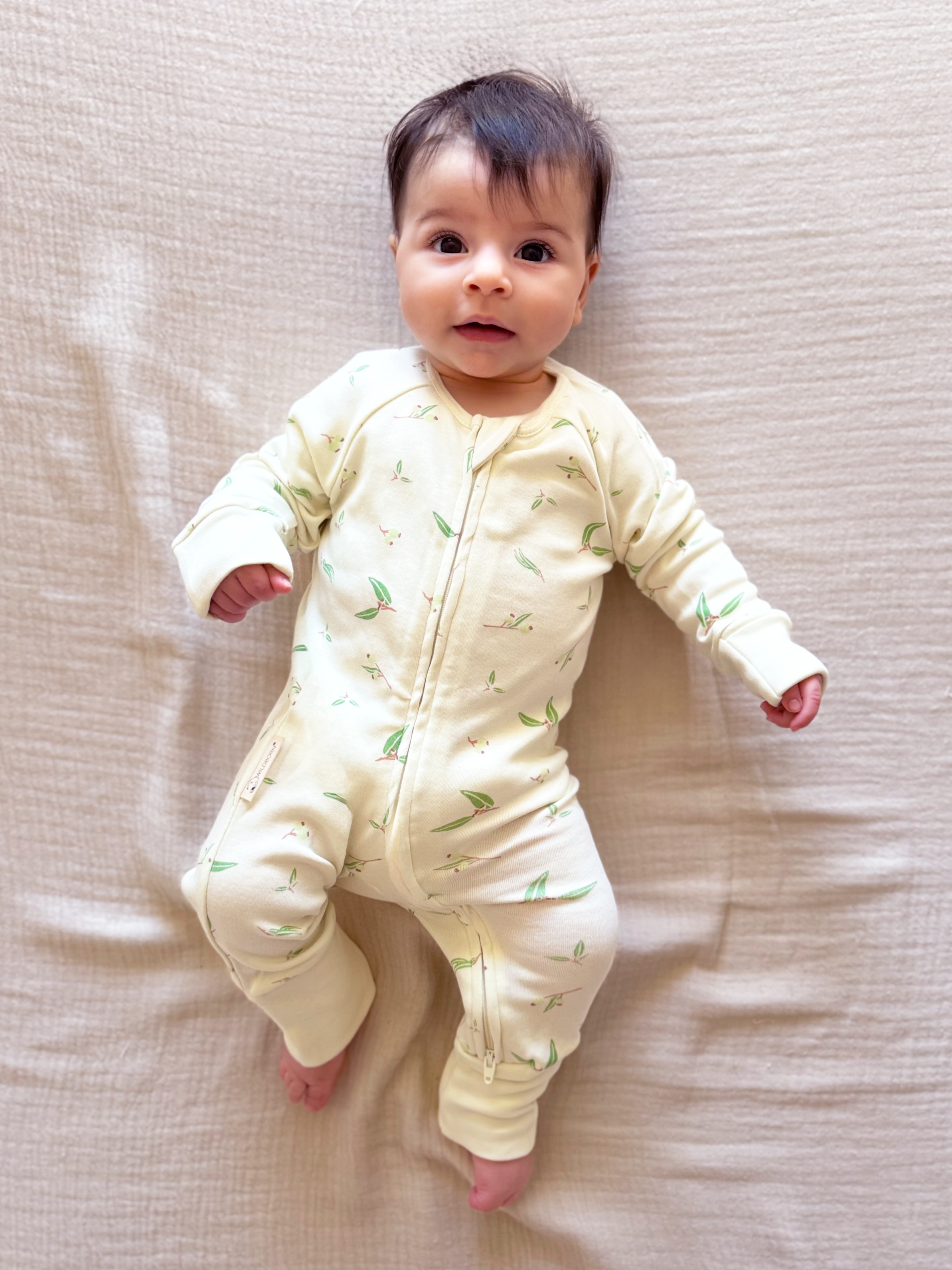 Baby wearing a light-colored onesie with a subtle pattern, lying on a textured beige surface.