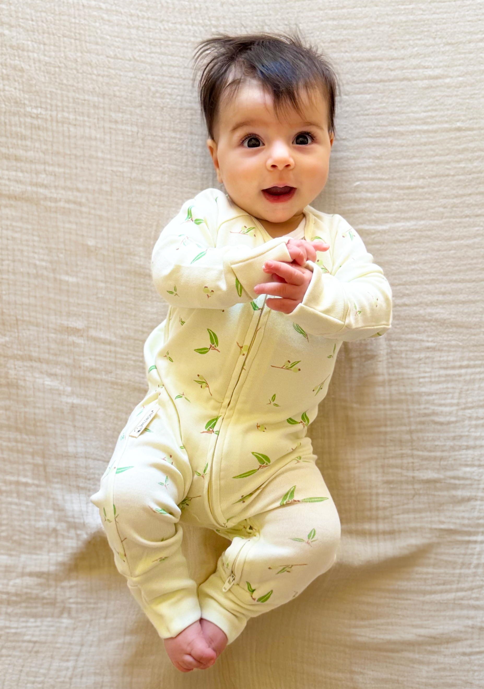 Baby wearing a yellow onesie with green patterns, lying on a beige surface.