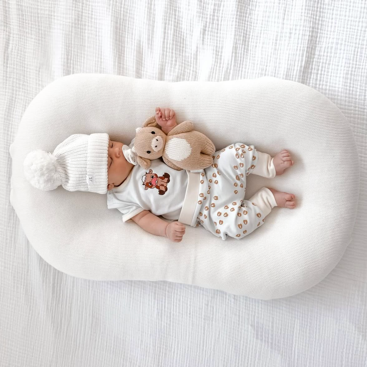 Baby lying on a white cushion with a cow toy, wearing a white beanie and outfit. 